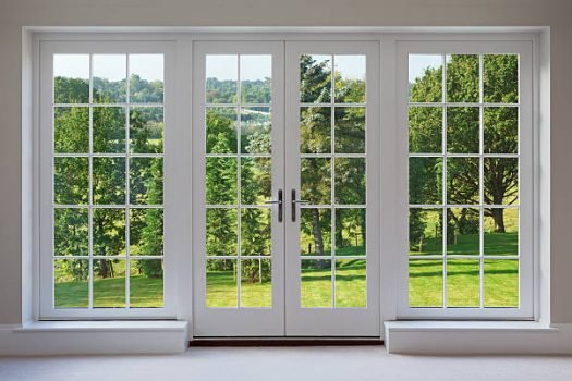 a set of finely crafted wooden Georgian style windows with doors set in a white frame, with white sills and caramel coloured walls. The view through the windows are towards a beautiful  countryside garden.